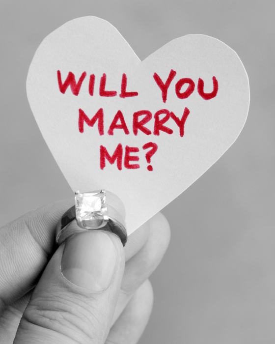 A hand wearing an engagement ring and holding a sign that says "Will you marry me?".